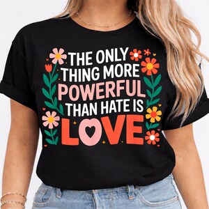 Può includere: T-shirt nero con la scritta "THE ONLY THING MORE POWERFUL THAN HATE IS LOVE" in bianco, rosa e arancione. Il design include elementi floreali rosa, arancioni e gialli, con foglie verdi. Una t-shirt grafica casual.