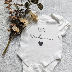 Könnte beinhalten: Weißer Baby Strampler mit schwarzem Text "MINI Nachname" und einem kleinen schwarzen Herz unter dem Text.