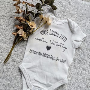 Könnte beinhalten: Weißer Baby-Strampler mit schwarzem Text, der "Alles Liebe zum ersten Valentinstag. Ich habe den tollsten Papa der Welt!" lautet. Der Strampler hat ein kleines schwarzes Herz in der Mitte des Textes.