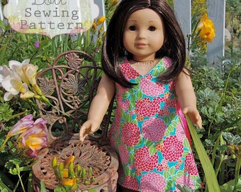 May include: A doll wearing a pink and green floral print dress with a V-neckline. The doll has long brown hair and is sitting on a metal chair in a garden setting.