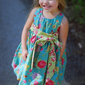 May include: A young girl wearing a blue sleeveless dress with a floral print and a green and yellow sash tied in a bow at the waist. The dress has a gathered skirt and a round neckline.