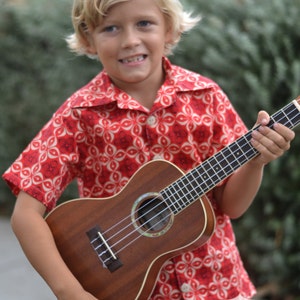 May include: A young person wearing a red and white patterned shirt is holding a brown ukulele. The shirt has a button-up collar and short sleeves.