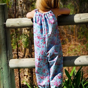 May include: A young girl wearing a blue and pink floral print jumpsuit stands on a wooden fence. She is looking off to the side, with her back to the camera.