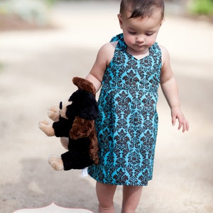 May include: A young child wearing a blue and black patterned sleeveless dress holds a brown and black stuffed animal. The child is standing on a sandy surface.