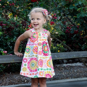 May include: A young girl wearing a white dress with a colorful floral pattern. The dress has a scoop neck and a gathered skirt. She is smiling and looking to the right. She is wearing a flower hair clip and sandals.