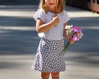 May include: A young girl wearing a white t-shirt and a gray and white polka dot skirt holds a white parasol and a bouquet of purple and white flowers. The girl is standing on a gray sidewalk.