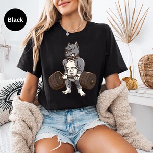 Puede incluir: Camiseta negra con un dibujo animado de un pitbull levantando una barra. El pitbull es gris y blanco, con una constitución musculosa y una expresión decidida. La barra es marrón. La palabra "Black" está en un círculo negro en la esquina superior izquierda.