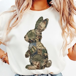 Può includere: Felpa color crema con un design a coniglio patchwork. Il coniglio è realizzato con vari tessuti mimetici, tra cui toppe verdi, marroni e blu. Il design è centrato sulla parte anteriore della maglietta, creando un'estetica unica e rustica.