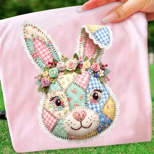 May include: A pink fabric item featuring a patchwork bunny face with floral accents. The bunny has a patchwork design with pink, blue, and yellow fabrics. The bunny's face is adorned with a floral crown and lace trim.