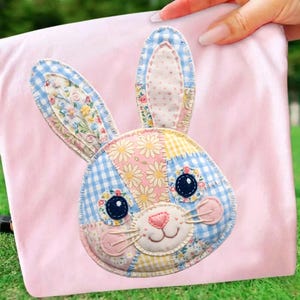May include: A pink fabric with a patchwork bunny face applique. The bunny has blue gingham, floral, and dotted fabric details. The bunny's eyes are dark blue, and the nose is pink.