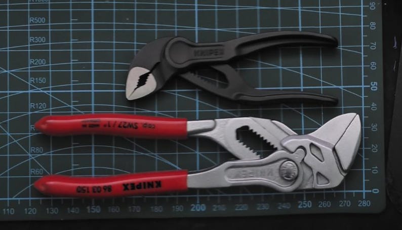 May include: Two pairs of pliers, one black and one silver, with red handles. The black pliers are labeled "KNIPEX" and the silver pliers are labeled "KNIPEX 86 03 150" and "CD SW27/1".