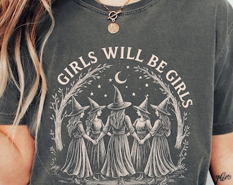 May include: A charcoal gray t-shirt featuring a cream-colored graphic of five figures in pointed hats holding hands in a circle under a crescent moon and stars. The text "GIRLS WILL BE GIRLS" is arched above the illustration, which is framed by trees.