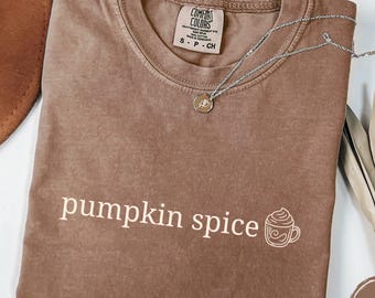 May include: A brown short-sleeved t-shirt with a crew neckline and the text "pumpkin spice" printed in white, accompanied by a small white line-art graphic of a coffee mug with whipped cream. The shirt features a Comfort Colors brand label.