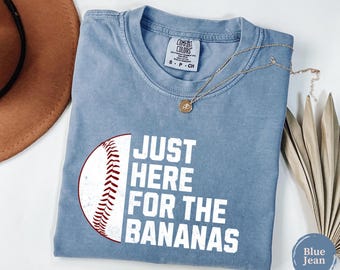 May include: A blue jean colored t-shirt featuring a white graphic of a baseball and the text "JUST HERE FOR THE BANANAS" in white block letters. The shirt has a crew neckline and a Comfort Colors brand label printed on the inside collar area.