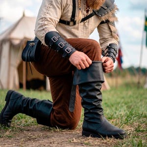 Peut inclure: Une personne à genoux ajuste une botte en cuir noir. Les bottes montent jusqu'aux genoux avec un revers. La personne porte un pantalon marron, une chemise crème et une armure de bras et d'épaules en cuir noir. Une sacoche en cuir est suspendue à la ceinture.