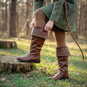 Pode incluir: Botas de couro marrom até o joelho com a parte superior dobrada. As botas têm uma textura ligeiramente enrugada e sola plana. A pessoa na imagem está usando meias marrons e uma túnica verde. Um arco é visível ao fundo.