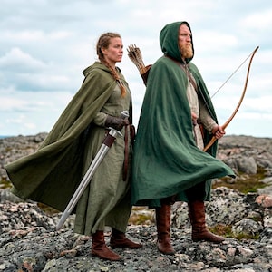 May include: Two people dressed in medieval attire stand on a rocky landscape. One holds a sword, the other a bow and arrow. Both wear hooded capes in shades of green and brown boots. The sky is overcast.