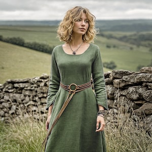 May include: A long-sleeved, olive green dress with a round neckline and a brown and gold belt. The dress is worn outdoors, with a stone wall and green hills in the background. The belt has a large gold ring buckle.