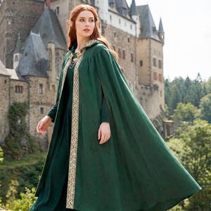 May include: A long, flowing green cloak with a hood and gold trim. The cloak is worn over a dark green dress. The background features a stone castle and trees, creating a fantasy or medieval theme.