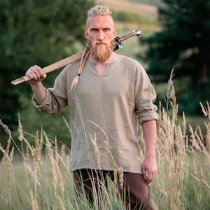 May include: A light brown linen tunic with a V-neck and long sleeves. The person is holding a sword with a wooden handle over their shoulder. The person has a long, braided beard and is standing in a field of tall grass.