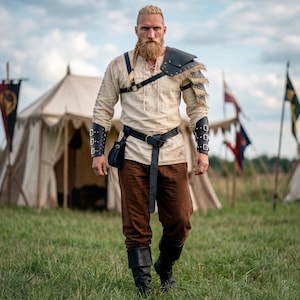 Pode incluir: Uma pessoa com uma roupa em estilo viking, incluindo uma túnica creme, calças castanhas e detalhes em couro preto. O conjunto apresenta uma ombreira, braçadeiras e um cinto com uma bolsa. A pessoa está andando em um campo gramado com tendas e bandeiras ao fundo.