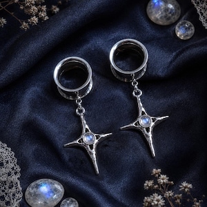 May include: A pair of silver-toned ear tunnels featuring dangling four-pointed star charms. Each charm has a textured, hammered metal surface and is set with a small, round, iridescent moonstone cabochon at the centre of the star design.