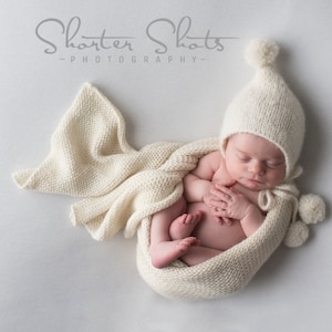 May include: A newborn baby wearing a white knitted hat with a pom-pom on top, swaddled in a white knitted blanket. The baby is sleeping peacefully with hands clasped together.
