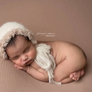 May include: A newborn baby wearing a white crocheted bonnet with a brown fur trim and a white crocheted scarf, sleeping on a brown textured background.