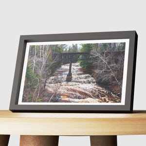 May include: A framed photograph of a wooden bridge over a rushing river in a forest. The bridge is made of dark wood and has a simple design. The river is flowing quickly and is surrounded by trees. The photo is taken from a low angle, looking up at the bridge.