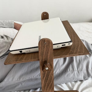 May include: A dark brown wooden laptop desk, supporting a white Samsung laptop. The desk is designed with a sloped surface for comfortable use in bed or on a sofa. The desk is made of wood.