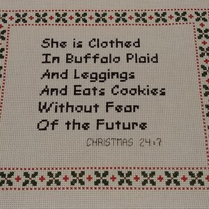 May include: Cross-stitch embroidery with a white background and a red and green border. The text reads "She is Clothed In Buffalo Plaid And Leggings And Eats Cookies Without Fear Of the Future. CHRISTMAS 24:7"
