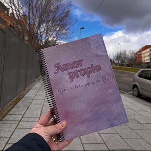 Op de afbeelding: Een spiraalgebonden notitieboek met een paars en roze aquarelontwerp. De omslag bevat de woorden "Amor Propio" en "Un ratito para mi" in een zacht, cursief lettertype. Het notitieboek wordt buiten tegen een bewolkte lucht gehouden.