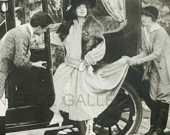 May include: Vintage black and white photograph of three people near a classic car. A woman in a dress and fur wrap exits the car, assisted by two people. The car has a visible steering wheel and a driver inside.