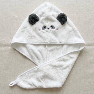 Op de afbeelding: Een witte handdoek met capuchon voor het haar, voorzien van een panda-gezicht met twee donkergrijze, textuurrijke oren, geborduurde zwarte ogen, een kleine neus en roze blosjes. De handdoek heeft een driehoekige vorm en een lusje.