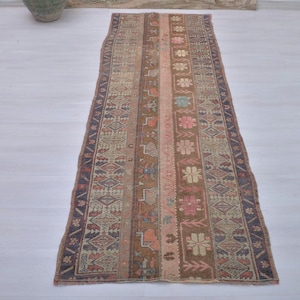 May include: A long, rectangular rug with a central design of vertical stripes in shades of brown, pink, and beige. The rug features intricate floral and geometric patterns, with a dark blue border. The rug is placed on a light-colored surface.