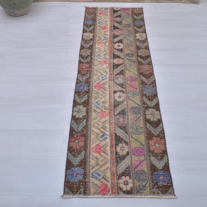 May include: A narrow, rectangular rug with a floral and geometric pattern. The rug features a central stripe with alternating floral and geometric designs in shades of brown, pink, blue, and green. The rug is placed on a light-colored wooden floor.