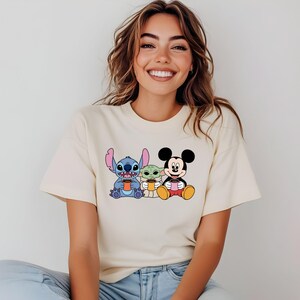 Puede incluir: Una camiseta de color crema con un gr&aacute;fico de Stitch, Baby Yoda y Mickey Mouse sosteniendo tazas.