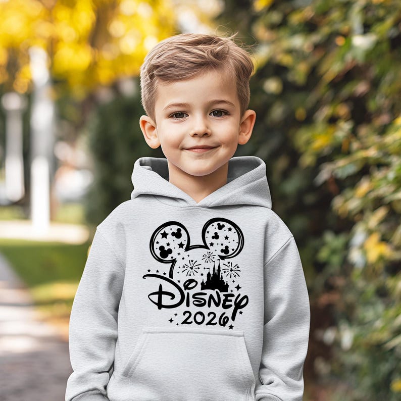 May include: A light gray hoodie featuring a black graphic of Mickey Mouse ears, fireworks, and a castle, with the text "Disney 2026". The hoodie is worn by a young child.