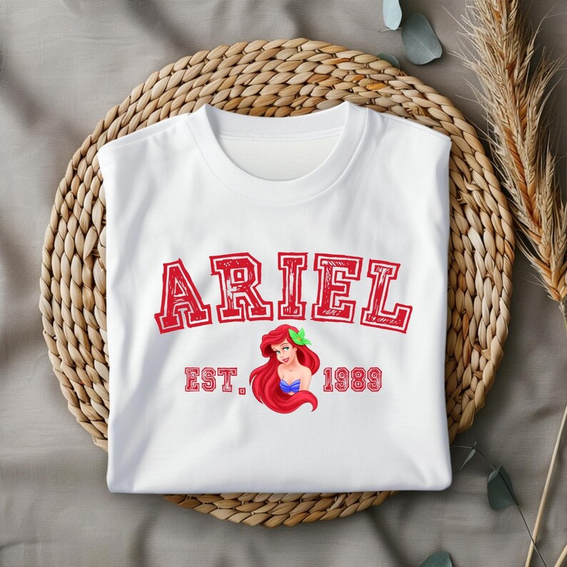 Puede incluir: Camiseta blanca con un gr&aacute;fico rojo de Ariel de La Sirenita. El gr&aacute;fico incluye el texto "ARIEL EST. 1989".