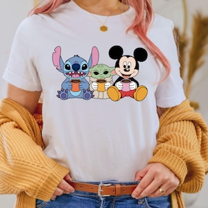 Puede incluir: Camiseta blanca con un gr&aacute;fico de Stitch, Baby Yoda y Mickey Mouse sosteniendo tazas de caf&eacute;.