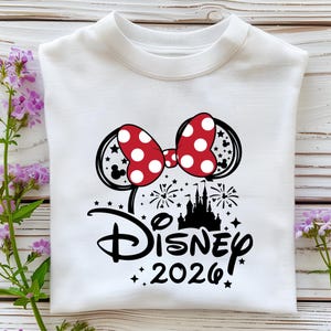 May include: White sweatshirt featuring a Minnie Mouse-themed design. The design includes a red bow with white polka dots, Mickey Mouse ears, a castle silhouette, fireworks, and the word "Disney" with the year "2026" in black.