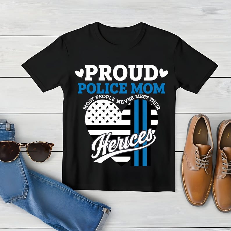 May include: Black t-shirt with a white and blue heart design. The heart features an American flag and a thin blue line. The text "Proud Police Mom" is above the heart and "Most People Never Meet Their Heros" is below the heart.