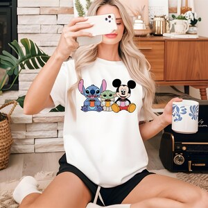 Puede incluir: Camiseta blanca con una imagen de dibujos animados de Stitch, Baby Yoda y Mickey Mouse sosteniendo tazas de caf&eacute;.