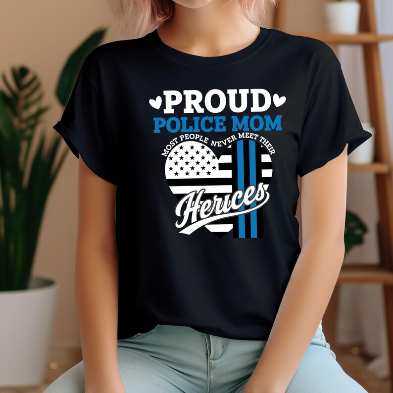 May include: Black t-shirt with a blue and white heart design. The heart features a thin blue line and an American flag. The text "Proud Police Mom" and "Most People Never Meet Their Heros" is printed on the shirt.