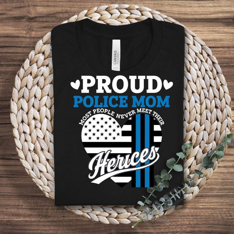 May include: Black t-shirt with a white heart design. The heart is divided in half, one side is the American flag and the other side is a blue and white police stripe. The text "Proud Police Mom" is above the heart and the text "Most people never meet their Heros" is below the heart.