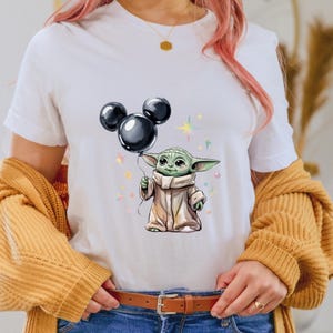 Op de afbeelding: Wit T-shirt met een groen personage dat een zwarte Mickey Mouse-ballon vasthoudt. Het personage draagt een bruine robe en heeft grote oren. De achtergrond is wit met kleurrijke sterren.