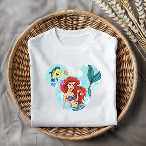 Puede incluir: Camiseta blanca con una imagen de dibujos animados de Ariel de La Sirenita, Flounder y una silueta de oreja de Mickey. La imagen est&aacute; en un esquema de color azul y verde.