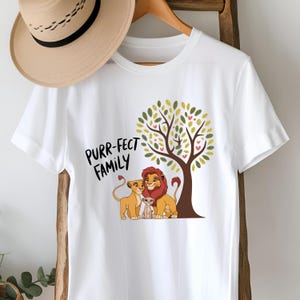 May include: White t-shirt with a graphic of a lion family under a tree with green leaves and pink hearts. The text "Purr-fect Family" is printed on the shirt.