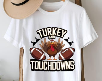 Könnte beinhalten: Weißes T-Shirt mit einem Cartoon-Truthahn und zwei American-Football-Bällen. Der Text "TURKEY AND TOUCHDOWNS" ist oberhalb und unterhalb des Truthahns und der Fußbälle aufgedruckt. Das T-Shirt hängt an einem Holzgestell.