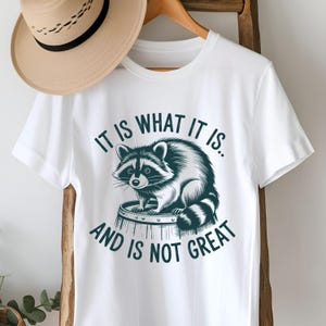 May include: White t-shirt with a graphic of a raccoon sitting on a barrel. The text on the shirt reads "It is what it is. And is not great."